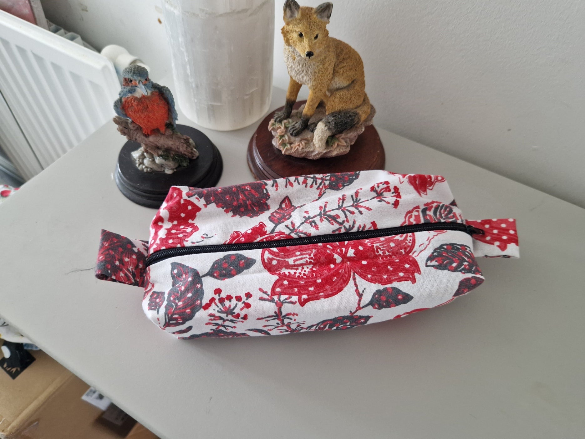 A fabric zippered pouch with a red and black floral/berry print on a white background, displayed on a light gray tabletop next to decorative figurines.