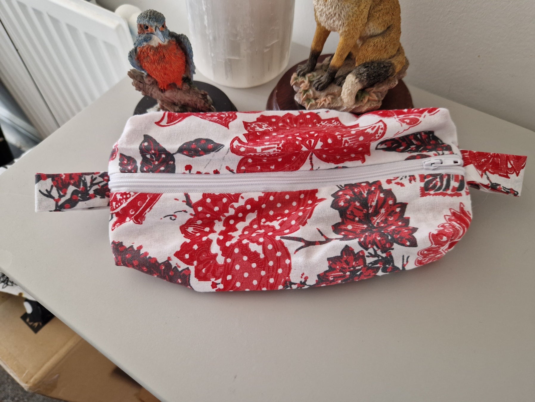 A fabric zippered pouch with a red and white floral print, displayed on a light-colored tabletop with decorative figurines in the background.