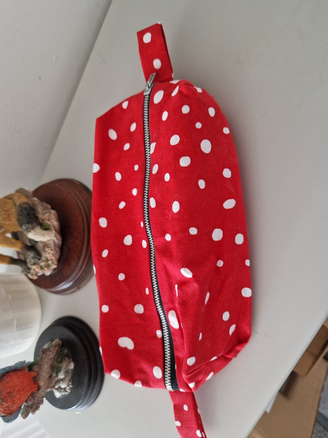 A red fabric zippered pouch with white polka dots, shown on a white tabletop with decorative objects in the background.