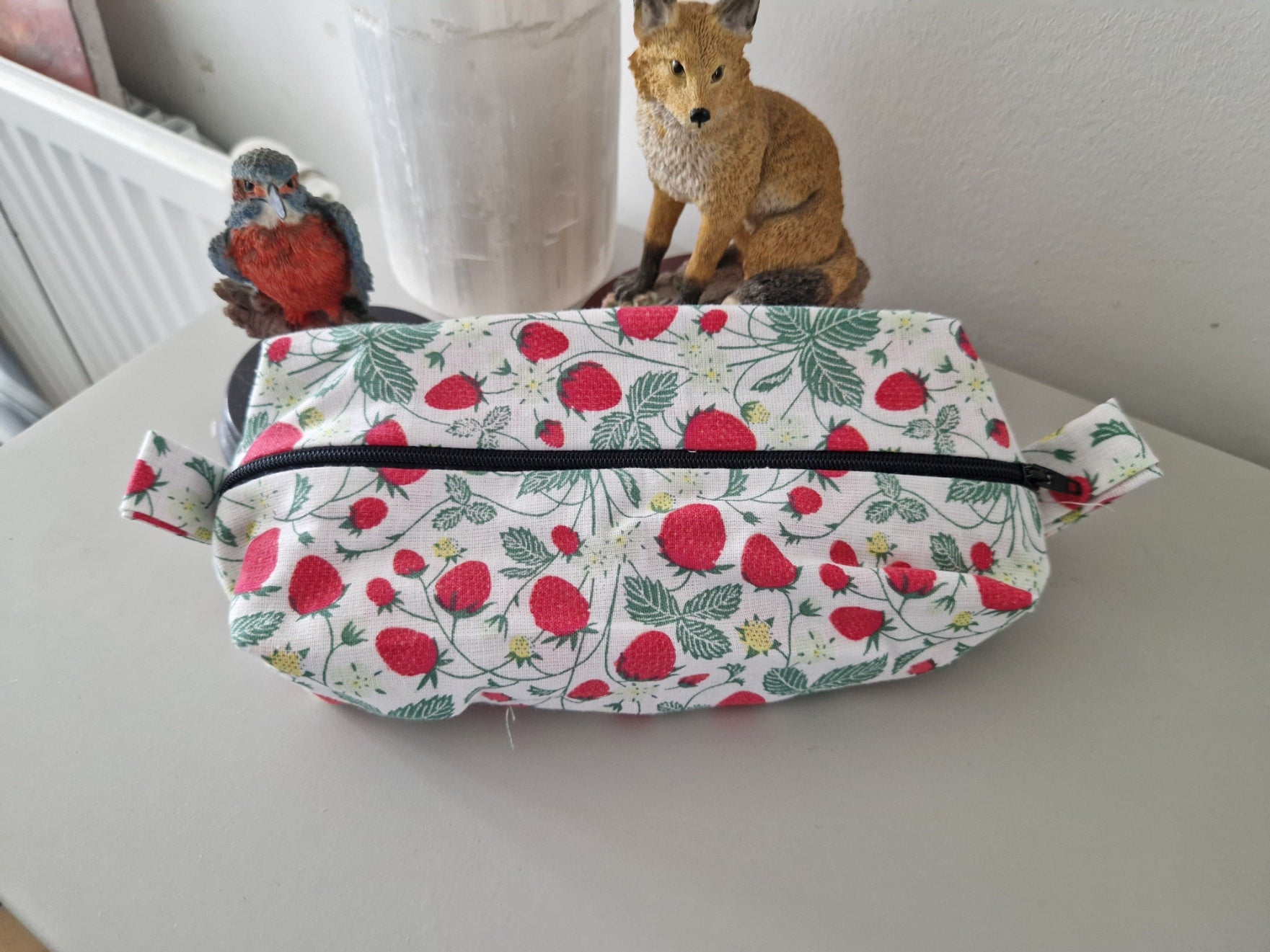 A fabric zippered pouch with a white background and a red strawberry and green leaf pattern, sitting on a light gray tabletop with decorative figurines behind it.