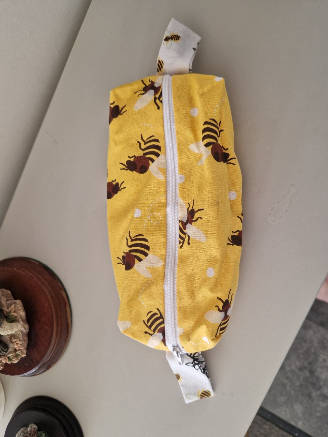 A yellow fabric zippered pouch decorated with a brown and black bee pattern, resting on a light gray tabletop.