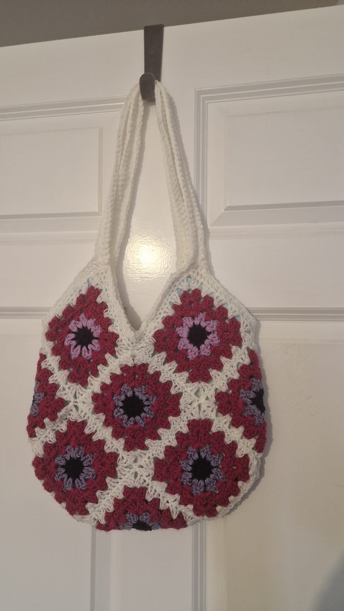 A handmade crocheted tote bag with white handles and a pattern of red, purple, and black granny squares, hanging on a dark hook against a white paneled door.