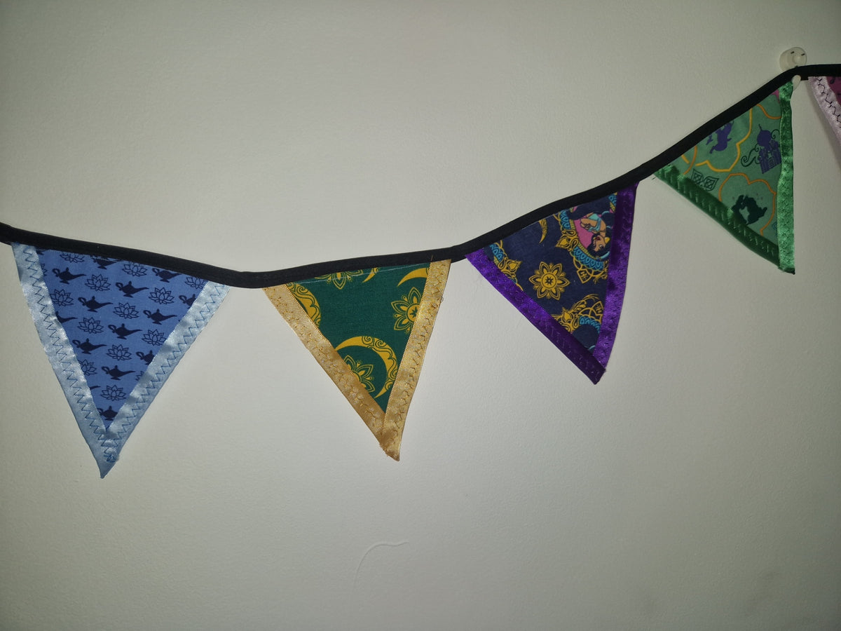 A fabric bunting banner with five triangular pennants hanging from a black cord against a plain off-white wall. The pennants feature different patterns and colors: light blue with black birds, dark green with gold borders and crescent moons, purple with gold celestial motifs, and green with gold borders.