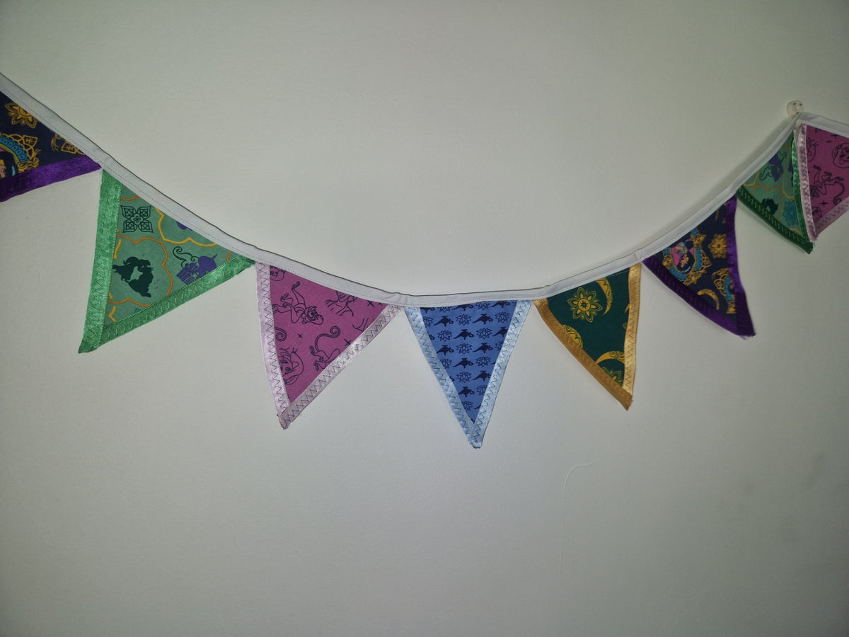 A string of triangular fabric bunting flags in assorted colors and patterns, hanging against a plain off-white wall.