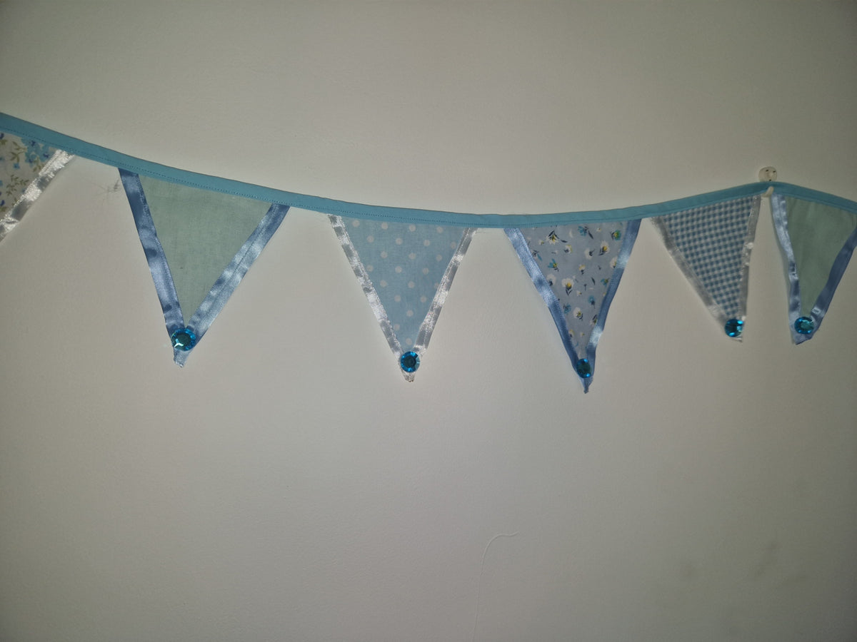 Light blue bunting banner with triangular flags in various patterns including polka dots, gingham, and stripes, hung against a plain off-white wall.