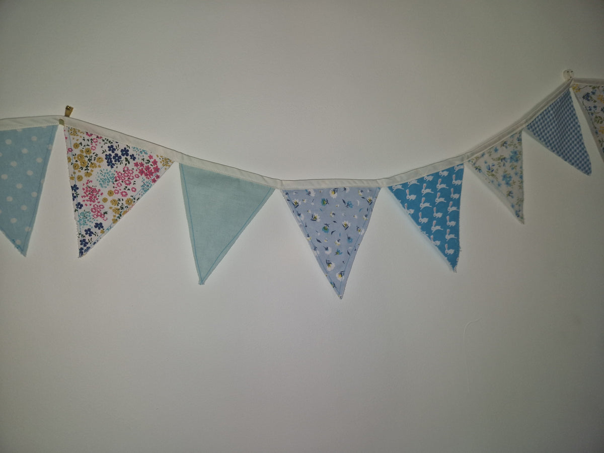 A fabric bunting banner with triangular flags in various blue patterns, including polka dots, floral prints, and geometric designs, hanging against a plain off-white wall.
