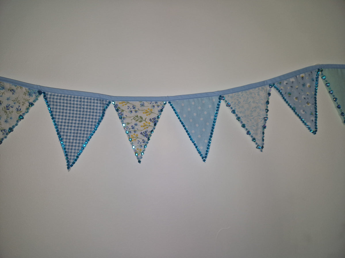 A light blue fabric bunting banner with triangular pennants in various patterns including gingham, polka dots, and floral prints, hung against a plain light gray wall.