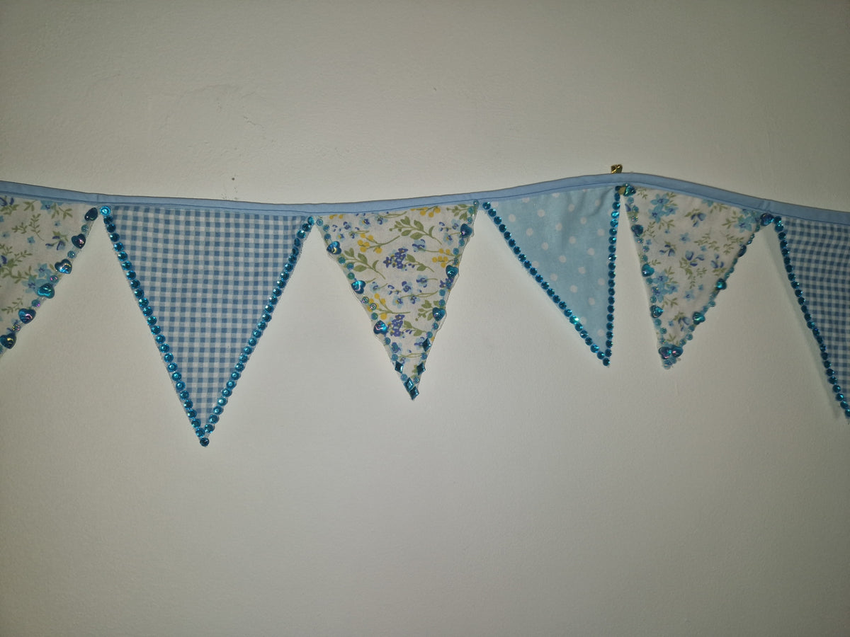 A fabric bunting banner with triangular flags in light blue gingham, light blue floral print, and light blue polka dot patterns, hanging against a plain off-white wall.