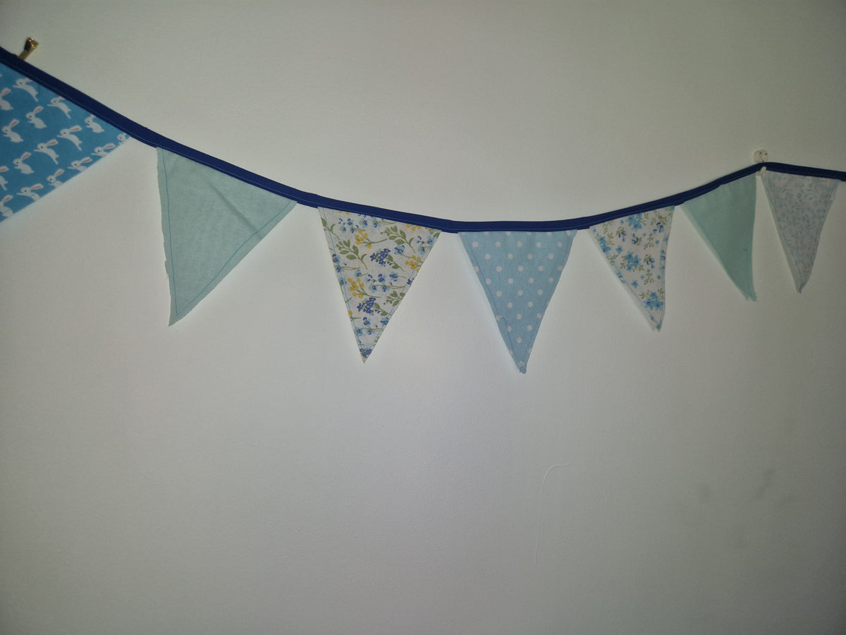 A string of triangular bunting flags in light blue, mint green, and floral patterns hanging against a plain off-white wall.