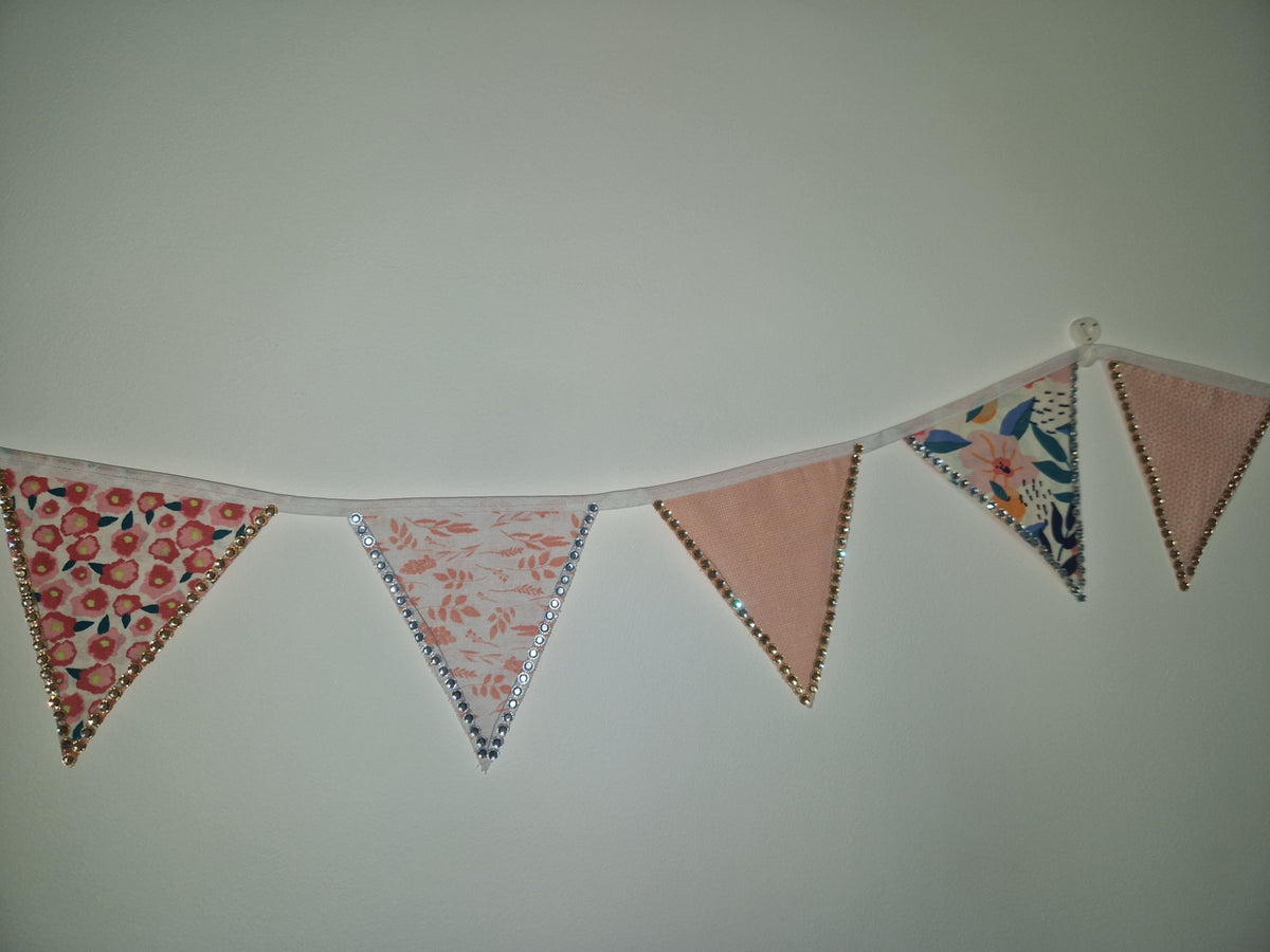A fabric bunting banner with six triangular flags hanging on a string against a plain light gray wall. The flags feature various patterns including floral prints, pink floral designs, and solid dusty pink, finished with silver glitter trim.
