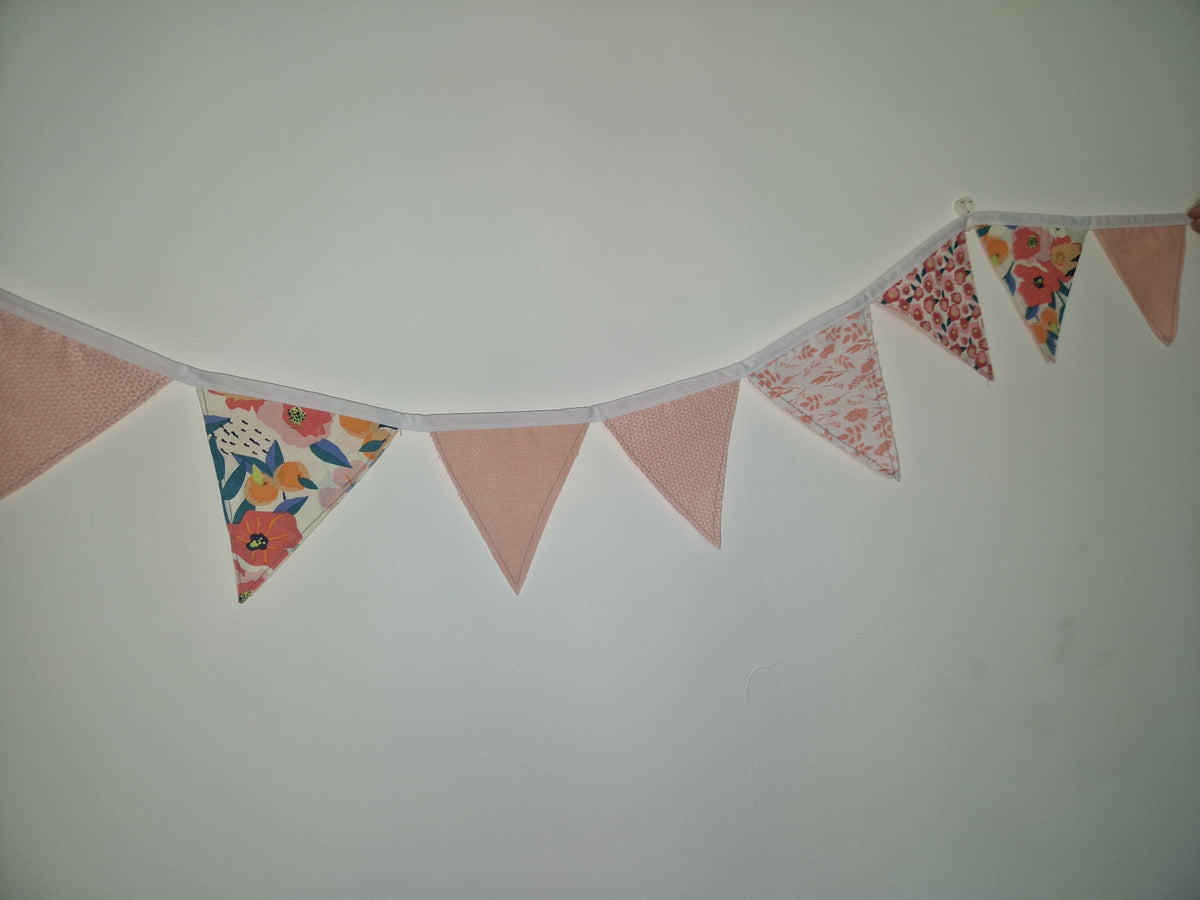 A string of triangular bunting flags hanging on a plain light wall. Several flags are solid dusty pink, while others have small floral and patterned prints in red, orange, blue, and cream.
