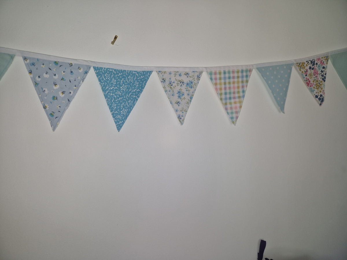 A string of triangular bunting flags in pastel blue, white, and multicolor floral/plaid patterns, hung against a plain white wall.