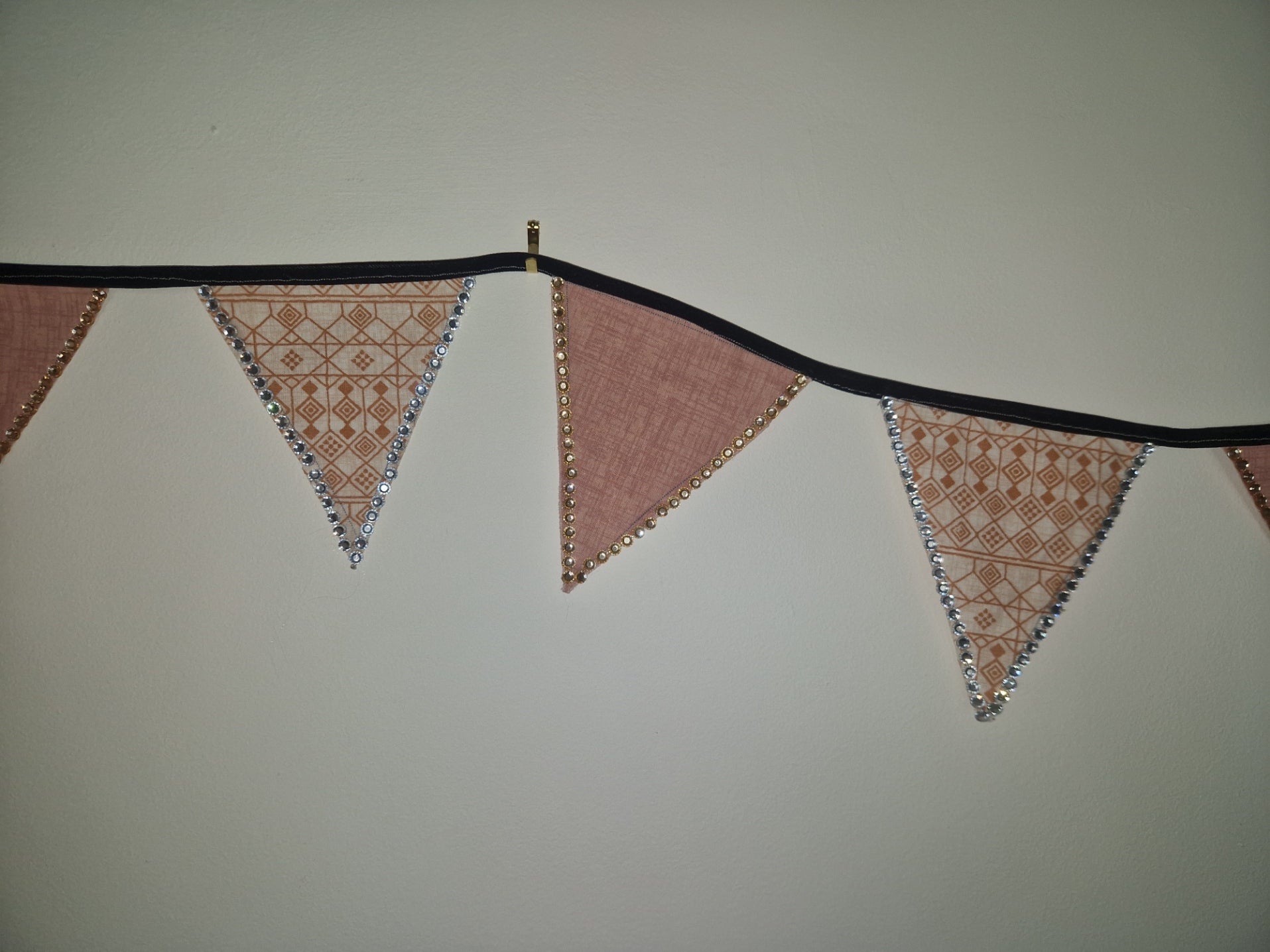 A rustic fabric bunting banner with alternating dusty pink and beige patterned pennants, strung along a black cord against a plain light gray wall.