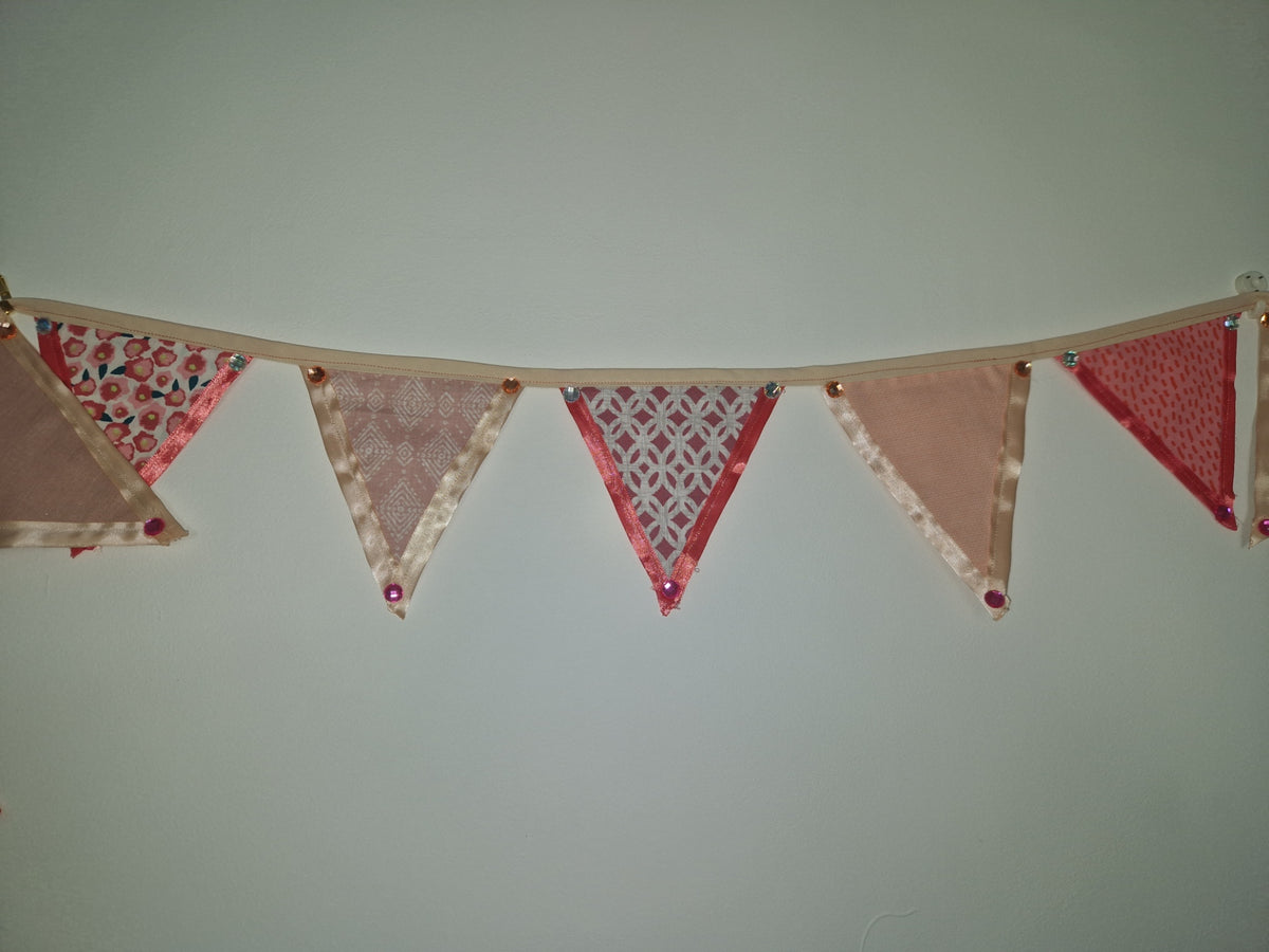 A handmade fabric bunting banner with five triangular flags in shades of pink, red, and beige with various patterns, strung along a wooden dowel against a plain light gray wall.