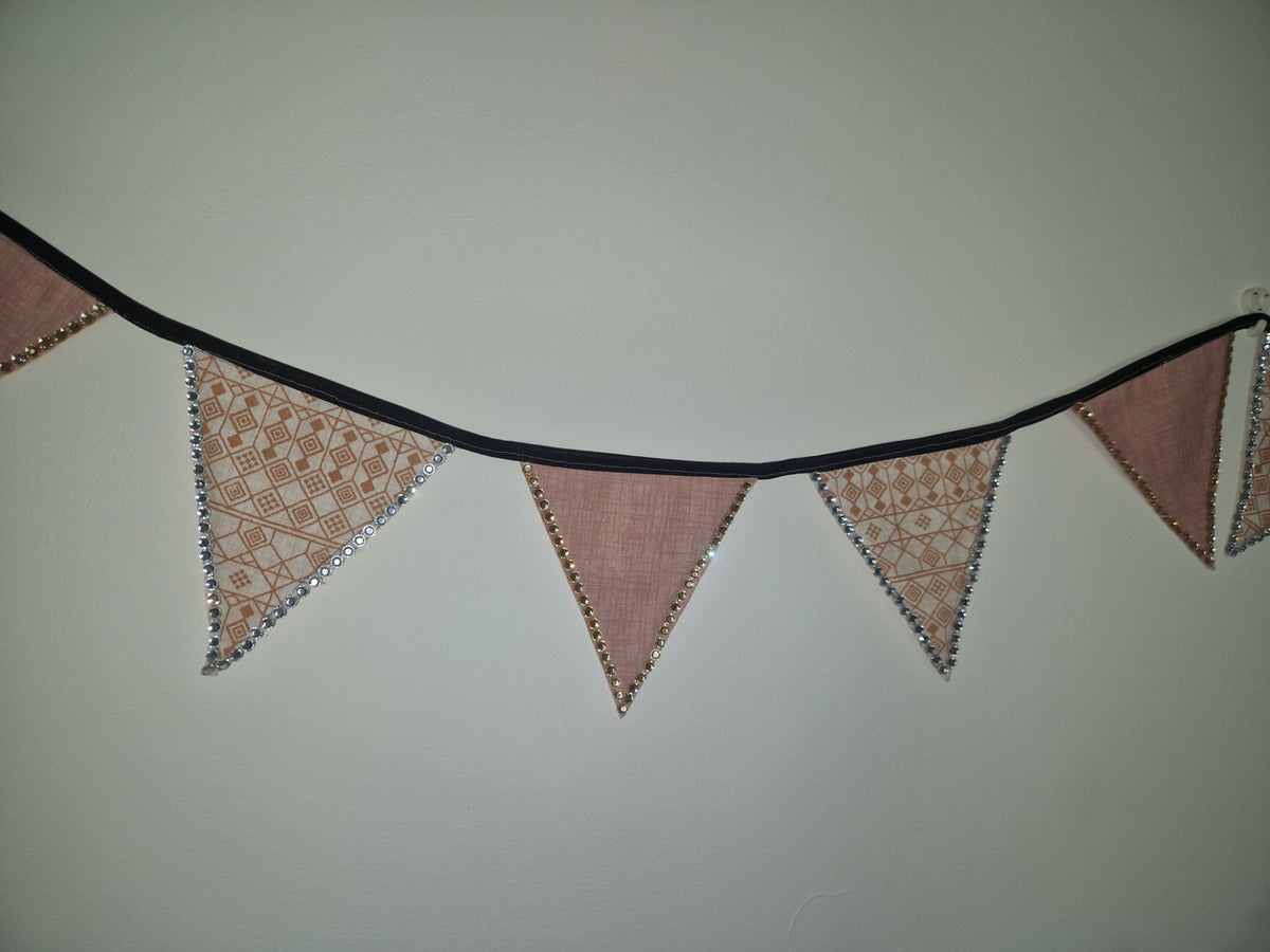 A rustic triangular bunting banner garland with dusty pink linen flags and silver embroidered borders, strung along a black cord against a plain light gray wall.