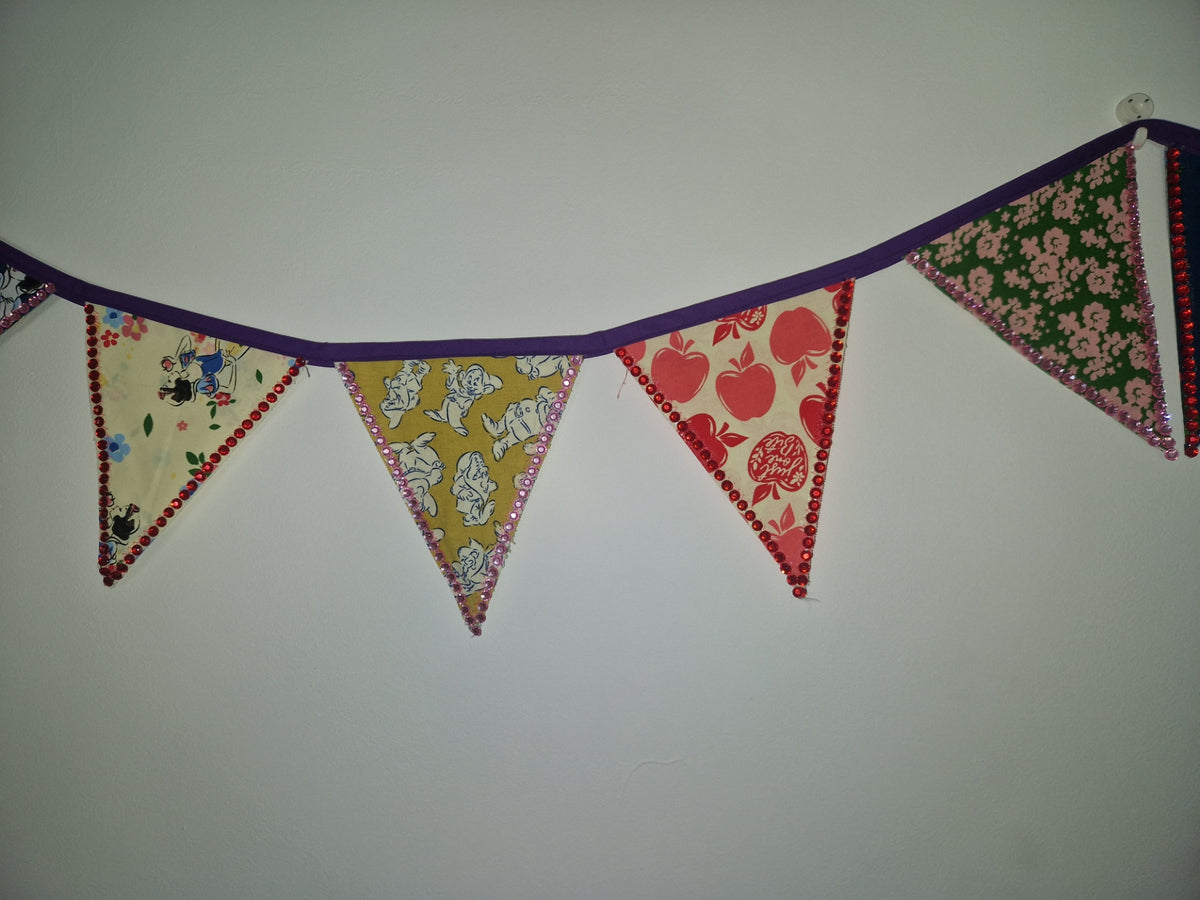 A fabric bunting banner with five triangular flags hanging on a purple cord against a plain light gray wall. The flags feature different patterns: a black and white cartoon print, a yellow and white animal print, a red and pink apple print, a green and pink floral print, and a red and white patterned edge.