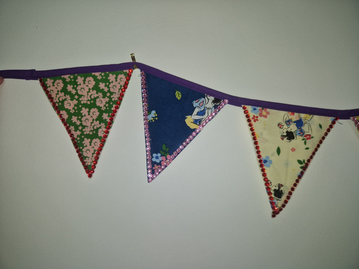 A fabric bunting banner with three triangular flags hanging from a purple cord. The left flag has pink flowers on a green background, the middle flag shows a blue pattern with a white character, and the right flag has a cream background with cartoon characters and flowers, all finished with red pom-pom trim.