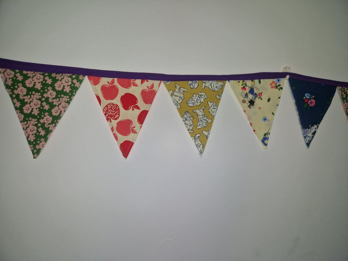 A fabric bunting banner with alternating triangular flags in floral and fruit patterns, hanging against a plain light gray wall.