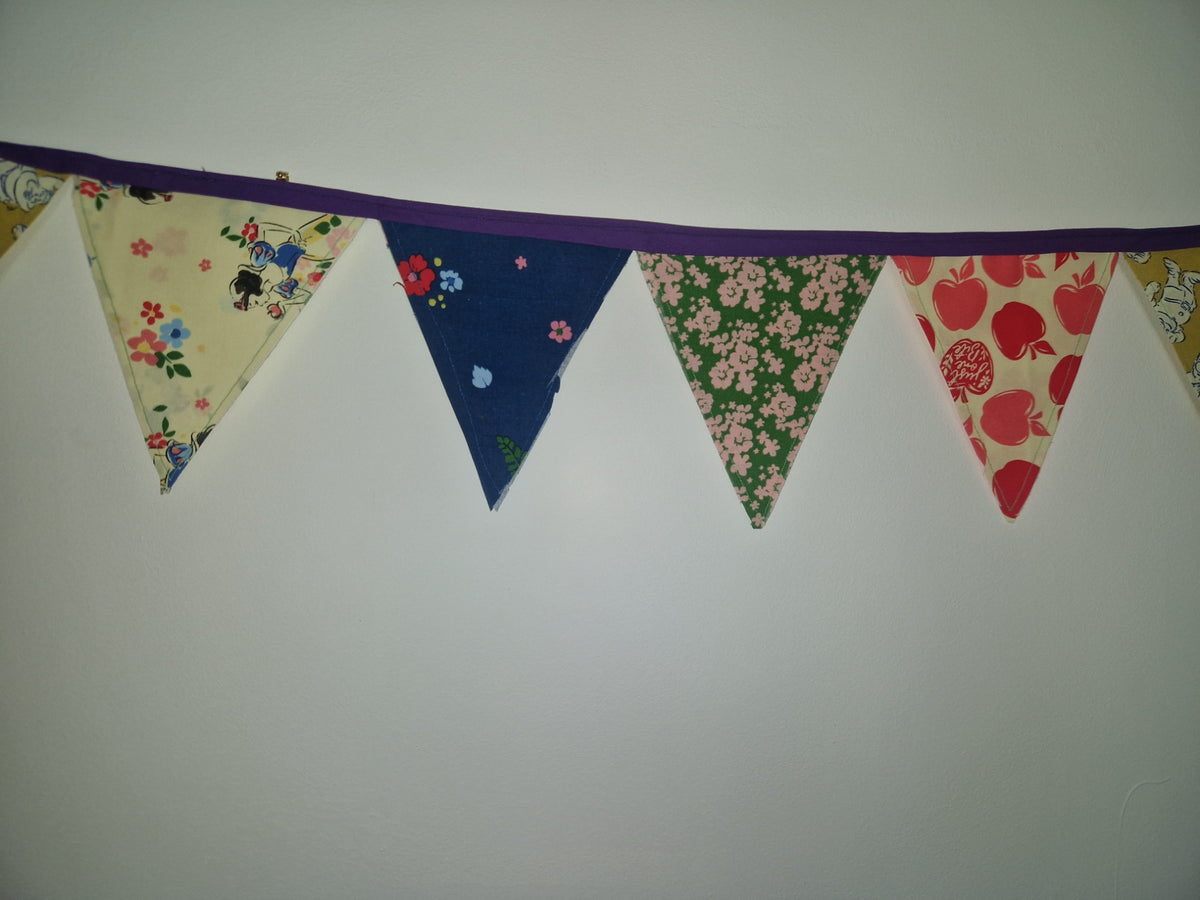 A fabric bunting banner with five triangular pennants hanging on a purple cord against a plain light gray wall. The pennants feature different patterns: a cream floral print, a dark blue floral print, a green floral print, a red apple print, and a beige patterned print.