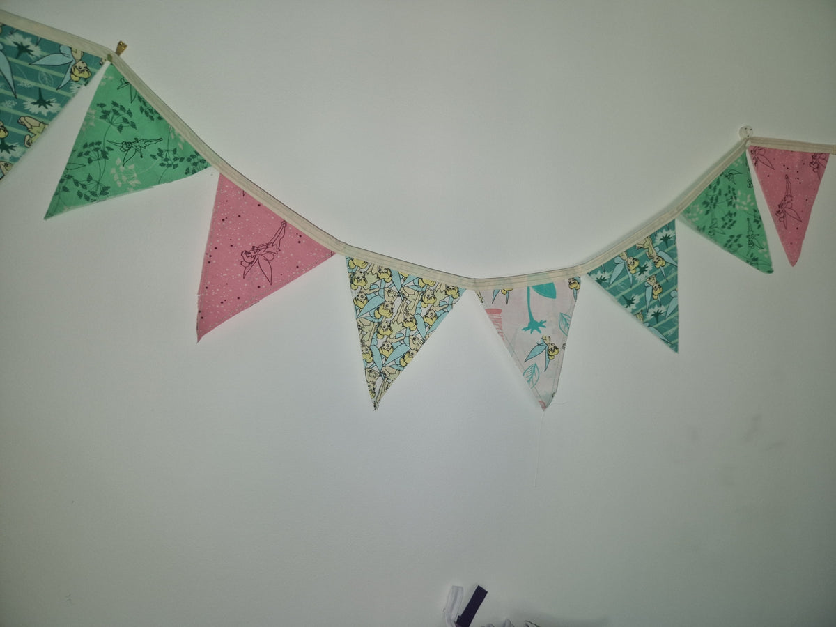 A fabric bunting banner with alternating triangular flags in green, pink, and light blue floral patterns, strung along a beige cord against a plain white wall.