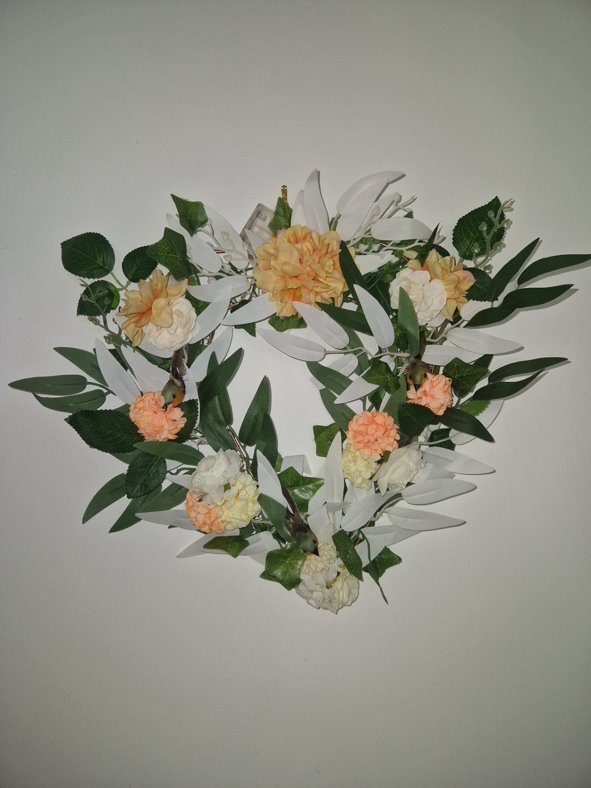 An artificial floral wreath featuring white lilies, peach roses, and green leaves, displayed against a plain light gray background.