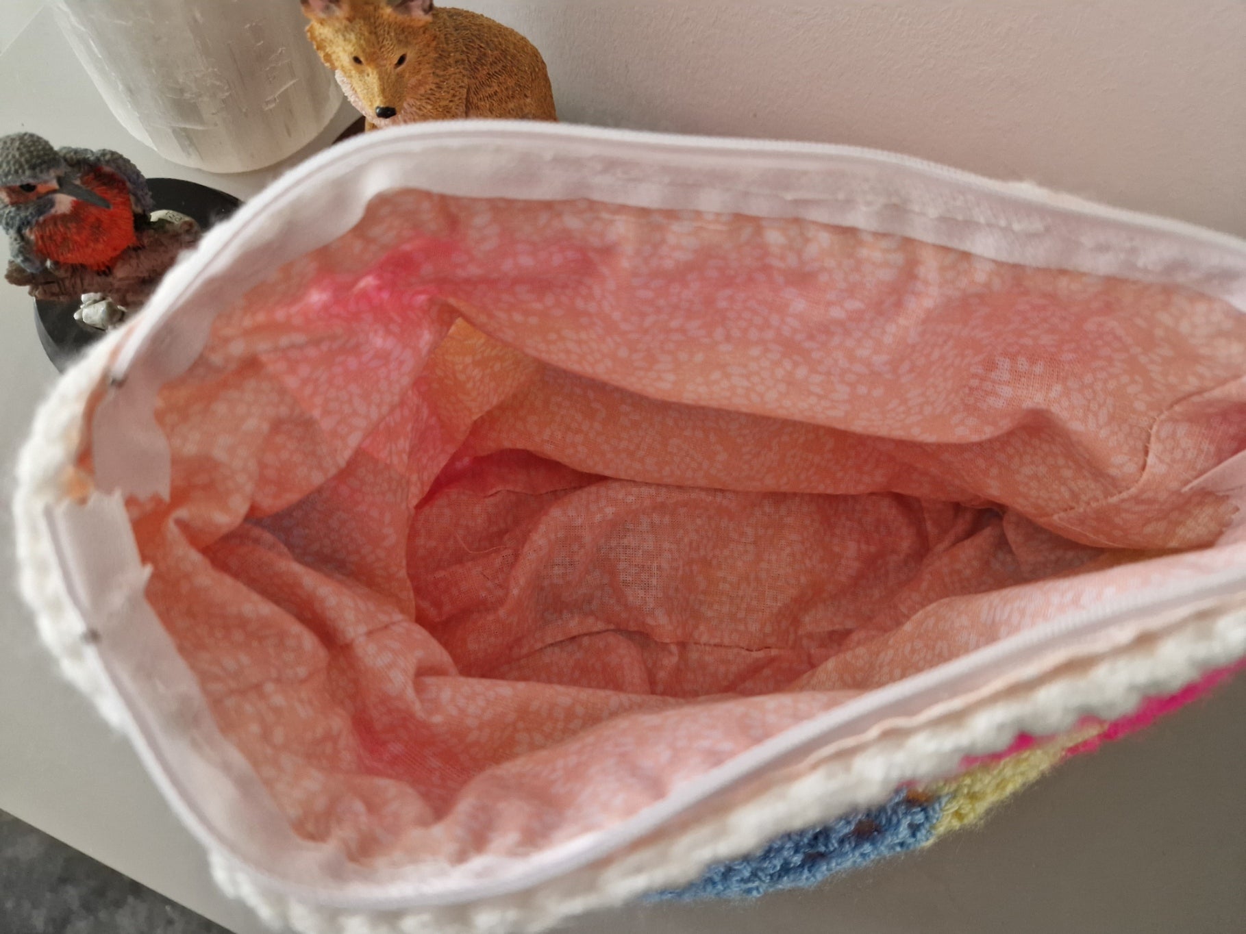 Close-up of the inside of a soft fabric pouch or bag with a pink and peach lining, white trim, and a small multicolored embroidered section visible at the bottom right.