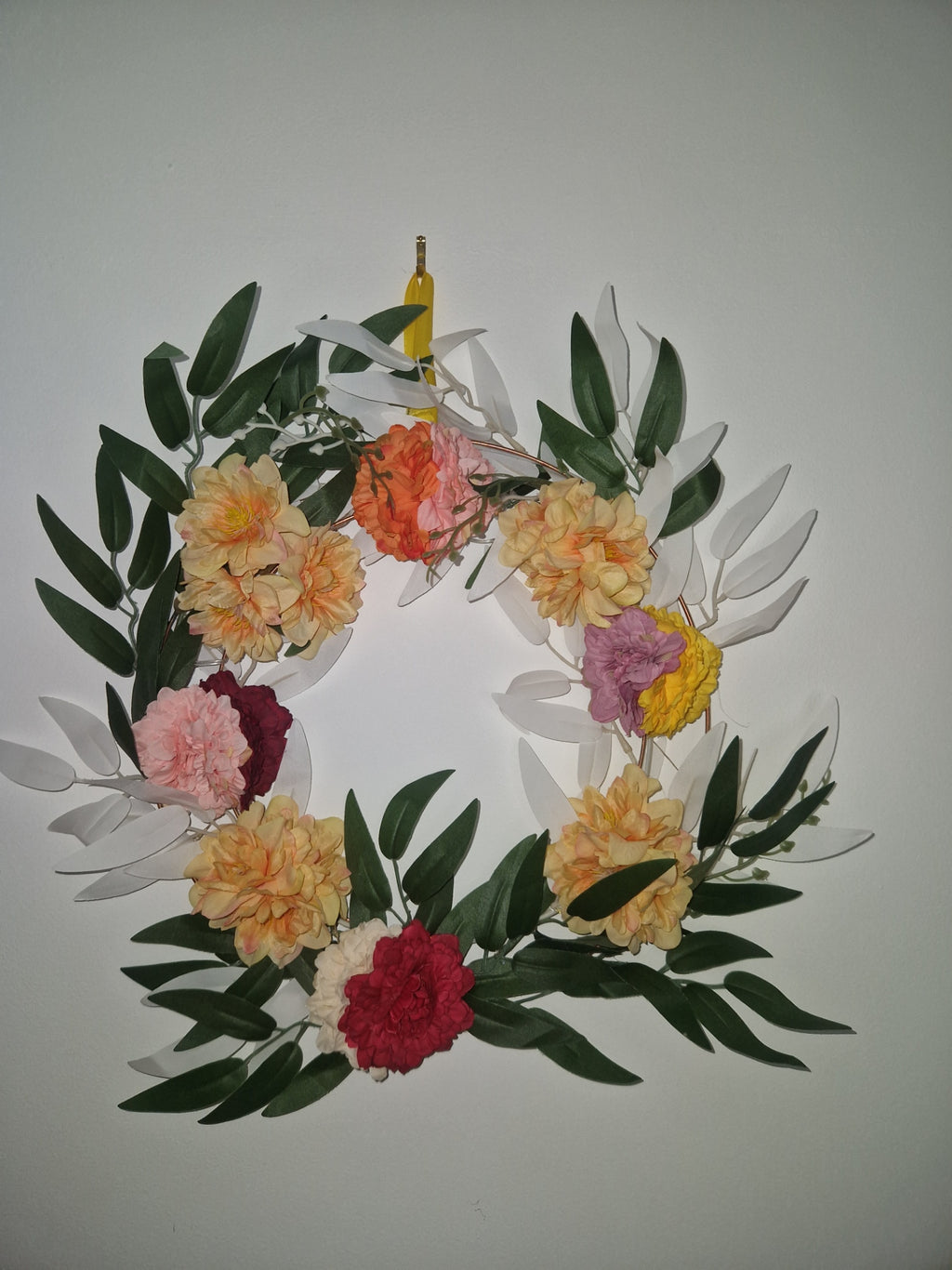 A floral wreath made of green leaves and artificial flowers in yellow, orange, pink, red, and purple, photographed against a plain light gray background.
