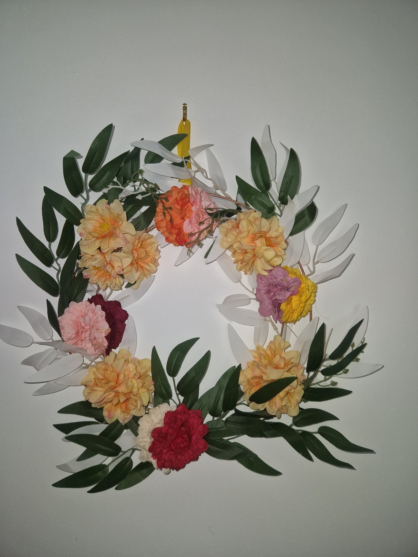 A floral wreath made of green leaves and artificial flowers in yellow, orange, pink, red, and purple, photographed against a plain light gray background.