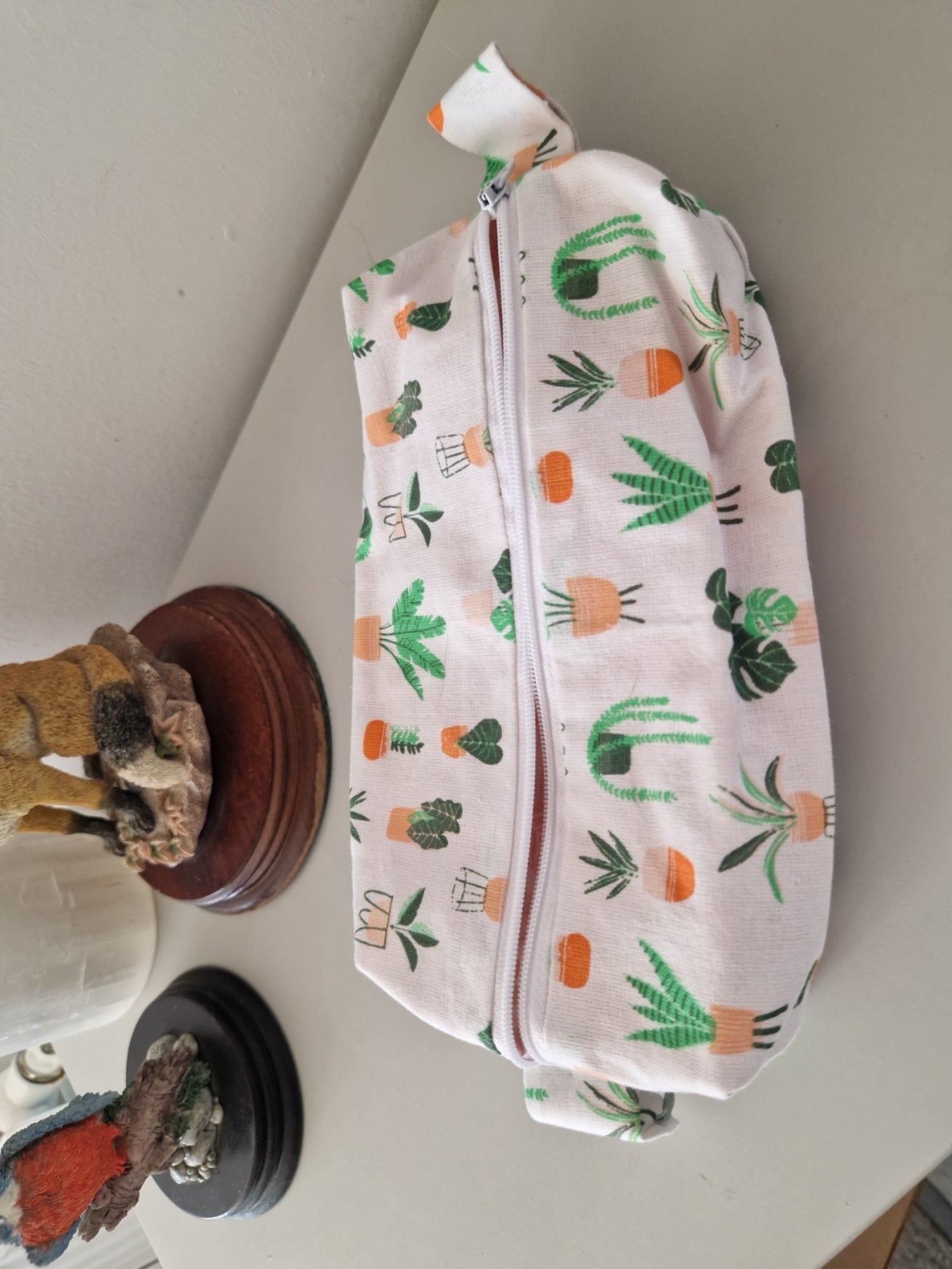 A fabric zippered pouch with a white background and a green, orange, and brown potted plant pattern, resting on a light gray tabletop with decorative figurines in the background.