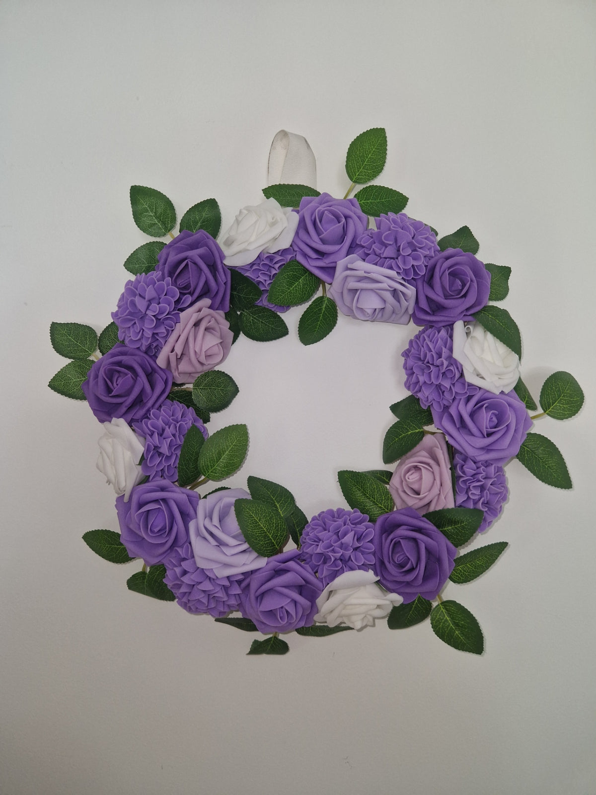 A decorative floral wreath made of purple and lavender fabric roses, hydrangeas, and green leaves, arranged in a circular shape against a plain light background.