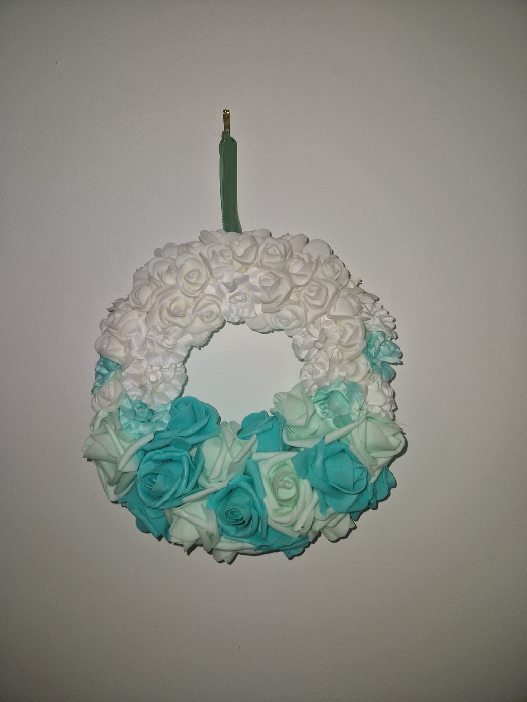 An artificial floral wreath with a gradient of white, mint green, and turquoise blue faux roses, attached to a green stem with a gold tip, shown against a plain light gray background.