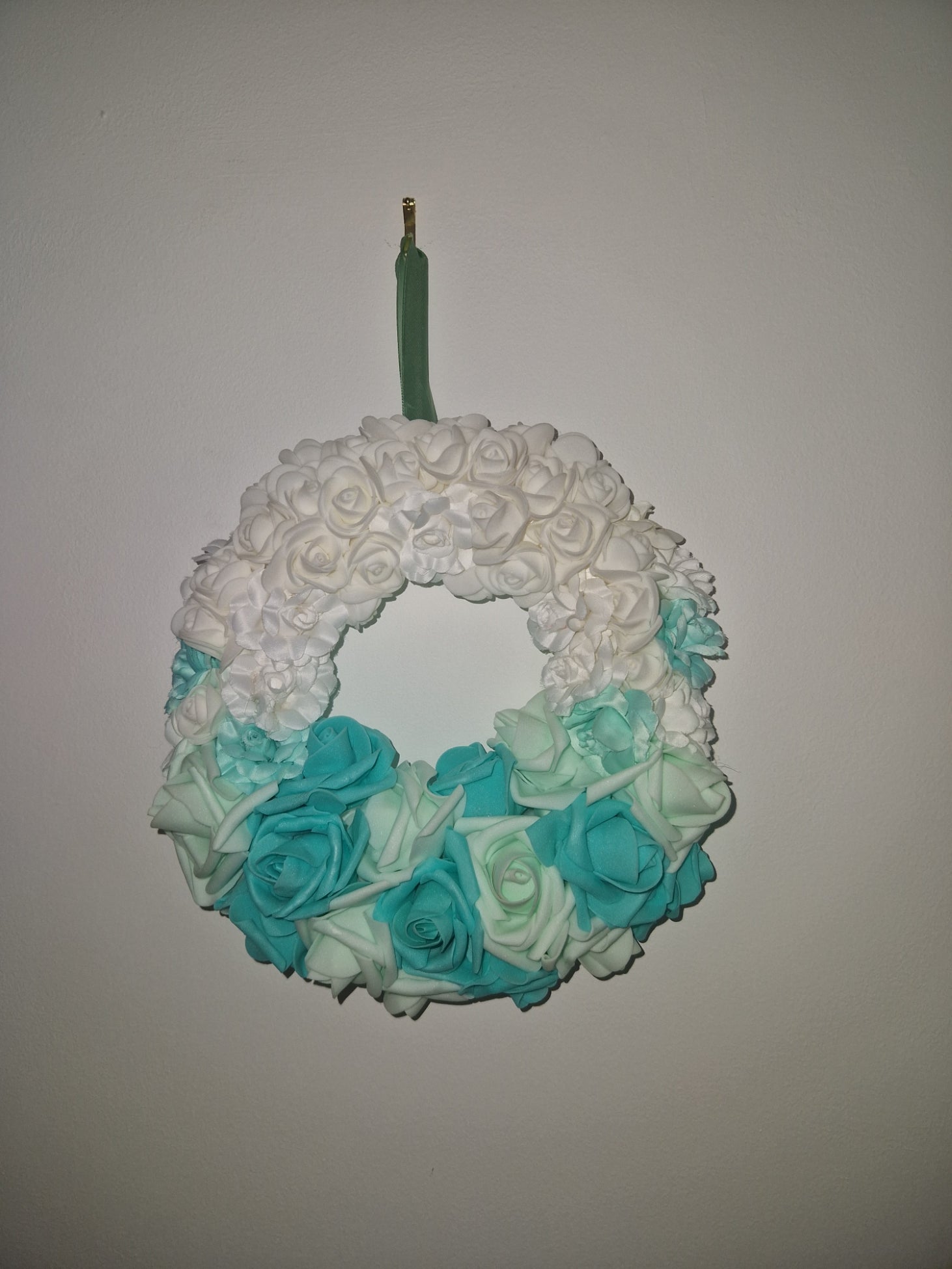 An artificial floral wreath with a gradient of white, mint green, and turquoise blue faux roses, attached to a green stem with a gold tip, shown against a plain light gray background.